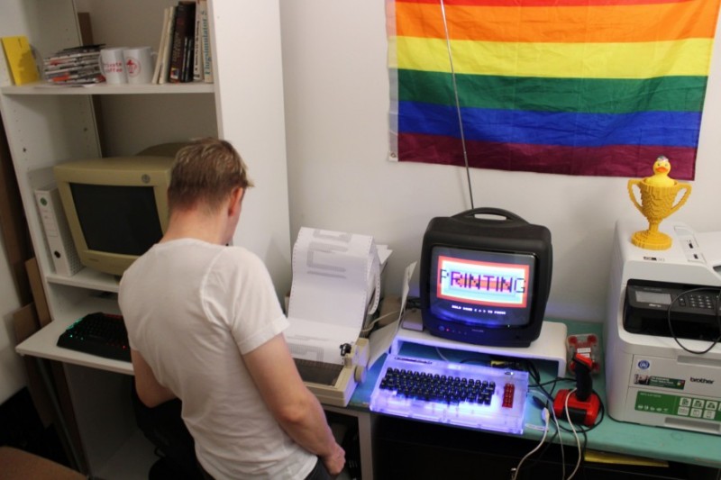 Picture of a person from the back playing around with a printer and fan-fold paper. To the left there is a CRT TV which reads Printing.