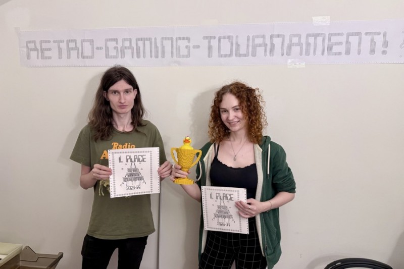 Picture of two people smilling at the camera. A banner above them reads Retro-Gaming-Tournament!. They hold up certificates reading "1st place". The person on the right is holding up a trophy with a duck inside.